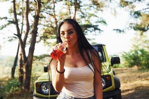 boire à la bouteille. blonde avec de l'alcool à la main se promener dans la forêt contre une jeep verte photo