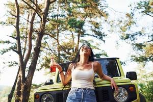 blonde avec de l'alcool à la main se promener dans la forêt contre la jeep verte photo