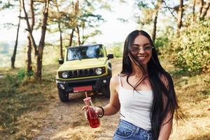 blonde avec de l'alcool à la main se promener dans la forêt contre la jeep verte photo