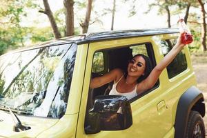 une fille avec une bouteille de boisson monte sa jeep verte moderne dans la forêt pendant la journée photo