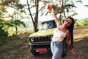 blonde avec de l'alcool à la main se promener dans la forêt contre la jeep verte photo