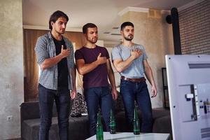 trois amis se tiennent à l'intérieur contre la télévision avec les mains sur le cœur. chant de l'hymne national photo