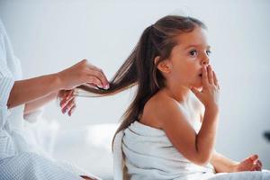 aider à faire la coiffure. jeune mère avec sa fille a une journée de beauté à l'intérieur dans une salle blanche photo