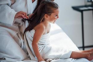 faire de la coiffure à l'aide d'un peigne. jeune mère avec sa fille a une journée de beauté à l'intérieur dans une salle blanche photo