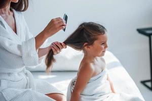 aider à faire la coiffure. jeune mère avec sa fille a une journée de beauté à l'intérieur dans une salle blanche photo