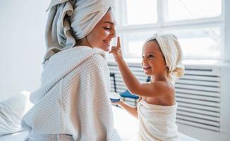 utiliser de la crème pour nettoyer la peau. jeune mère avec sa fille a une journée de beauté à l'intérieur dans une salle blanche photo