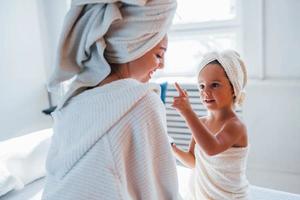 utiliser de la crème pour nettoyer la peau. jeune mère avec sa fille a une journée de beauté à l'intérieur dans une salle blanche photo