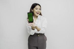 femme asiatique excitée portant une chemise blanche pointant vers l'espace de copie à côté d'elle tout en tenant son téléphone, isolée par fond blanc photo