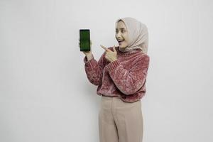 femme musulmane asiatique excitée portant un pull rose et un hijab pointant vers l'espace de copie à côté d'elle tout en tenant son téléphone, isolée sur fond blanc photo