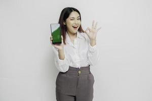 un portrait d'une femme asiatique excitée portant une chemise blanche tout en tenant son téléphone et en gesticulant signe d'approbation, isolée sur fond blanc photo