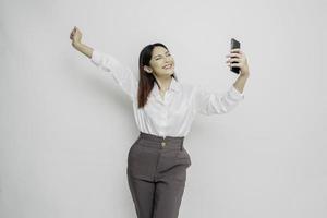 une jeune femme asiatique avec une expression heureuse et réussie portant une chemise blanche et tenant un smartphone isolé sur fond blanc photo