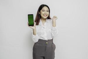 femme asiatique excitée portant une chemise blanche pointant vers l'espace de copie à côté d'elle tout en tenant son téléphone, isolée par fond blanc photo