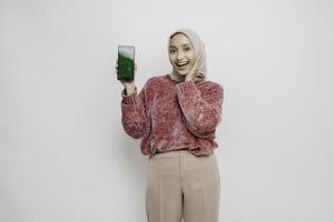 femme musulmane asiatique excitée portant un pull rose et un hijab pointant vers l'espace de copie à côté d'elle tout en tenant son téléphone, isolée sur fond blanc photo