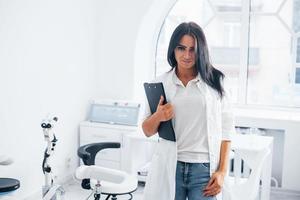 avec bloc-notes dans les mains. femme médecin brune se tient dans un bureau moderne et regarde la caméra photo