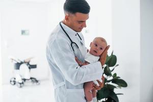 se dresse contre la fenêtre. le jeune pédiatre est avec un petit bébé à la clinique pendant la journée photo