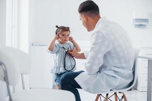 enseigne à la fille comment utiliser le stéthoscope. jeune pédiatre travaille avec une petite visiteuse à la clinique photo