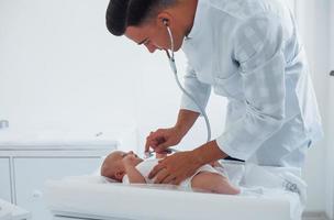 écoute à l'aide d'un stéthoscope. le jeune pédiatre est avec un petit bébé à la clinique pendant la journée photo