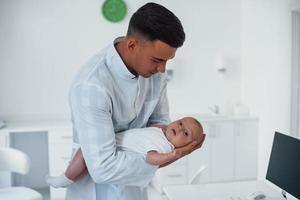 se tient debout et tient bébé sur les mains. jeune pédiatre est à la clinique pendant la journée photo