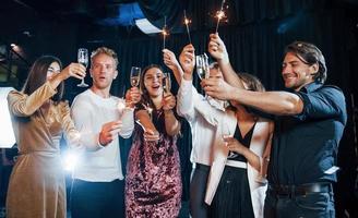 s'amuser avec des cierges magiques. groupe d'amis joyeux célébrant le nouvel an à l'intérieur avec des boissons dans les mains photo