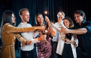 s'amuser avec des cierges magiques. groupe d'amis joyeux célébrant le nouvel an à l'intérieur avec des boissons dans les mains photo