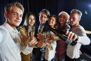 prend un selfie. groupe d'amis joyeux célébrant le nouvel an à l'intérieur avec des boissons dans les mains photo