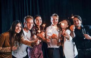 s'amuser avec des cierges magiques. groupe d'amis joyeux célébrant le nouvel an à l'intérieur avec des boissons dans les mains photo