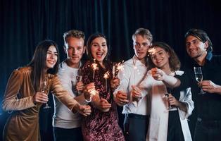 s'amuser avec des cierges magiques. groupe d'amis joyeux célébrant le nouvel an à l'intérieur avec des boissons dans les mains photo