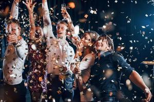 les confettis sont dans l'air. groupe d'amis joyeux célébrant le nouvel an à l'intérieur avec des boissons dans les mains photo