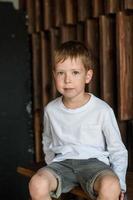 portrait d'un petit garçon blond en t-shirt blanc est assis sur le fond d'un mur en bois et sourit, regarde la caméra. photo
