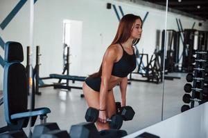 jeune femme fitness est dans la salle de gym près du miroir avec des haltères dans les mains photo