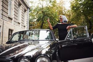 femme blonde à lunettes de soleil et en robe noire près de la vieille voiture classique vintage photo