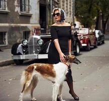 femme blonde à lunettes de soleil et en robe noire près de la vieille voiture classique vintage avec son chien photo