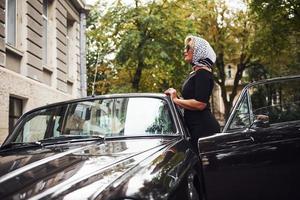 femme blonde à lunettes de soleil et en robe noire près de la vieille voiture classique vintage photo