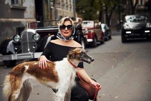 femme blonde à lunettes de soleil et en robe noire près de la vieille voiture classique vintage avec son chien photo