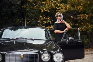 femme blonde à lunettes de soleil et en robe noire près de la vieille voiture classique vintage photo