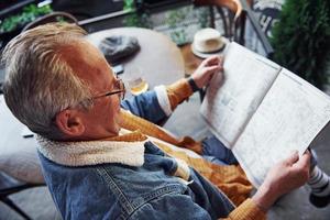 senior élégant dans des vêtements à la mode et des lunettes est assis dans le café et lit le journal