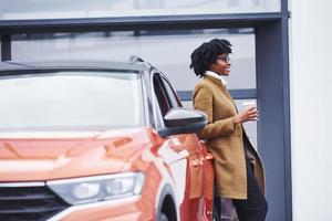 jeune femme afro-américaine dans des verres et avec une tasse de boisson se tient à l'extérieur près d'une voiture moderne photo