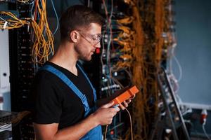 jeune homme en uniforme avec appareil de mesure fonctionne avec un équipement internet et des fils dans la salle des serveurs photo