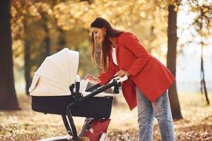 mère en manteau rouge se promener avec son enfant dans le landau dans le parc avec de beaux arbres à l'automne photo