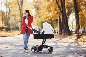 parler au téléphone. mère en manteau rouge se promener avec son enfant dans le landau dans le parc à l'automne photo