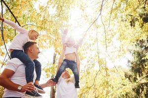 la mère et le père tiennent les enfants aux épaules et dans les mains. joyeuse jeune famille se promener ensemble dans un parc d'automne photo