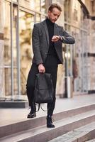 homme en tenue de soirée élégante et avec sac est à l'extérieur contre un bâtiment moderne photo