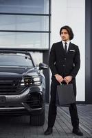 portrait d'un beau jeune homme d'affaires en costume noir et cravate à l'extérieur près d'une voiture moderne et avec des sacs à provisions photo