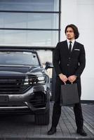 portrait d'un beau jeune homme d'affaires en costume noir et cravate à l'extérieur près d'une voiture moderne et avec des sacs à provisions photo