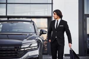 portrait d'un beau jeune homme d'affaires en costume noir et cravate à l'extérieur près d'une voiture moderne et avec des sacs à provisions photo