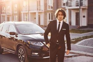 portrait d'un beau jeune homme d'affaires en costume noir et cravate à l'extérieur près d'une voiture moderne dans la ville photo