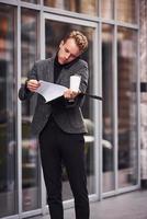 homme en tenue de soirée élégante avec une tasse de boisson et un bloc-notes dans les mains est à l'extérieur contre un bâtiment moderne avoir une conversation par téléphone photo