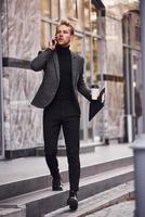 homme en tenue de soirée élégante avec une tasse de boisson et un bloc-notes dans les mains est à l'extérieur contre un bâtiment moderne avoir une conversation par téléphone photo