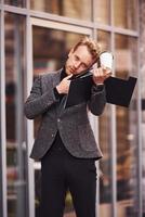 homme en tenue de soirée élégante avec une tasse de boisson et un bloc-notes dans les mains est à l'extérieur contre un bâtiment moderne avoir une conversation par téléphone photo