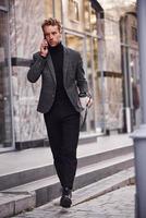 homme en tenue de soirée élégante avec une tasse de boisson et un bloc-notes dans les mains est à l'extérieur contre un bâtiment moderne avoir une conversation par téléphone photo